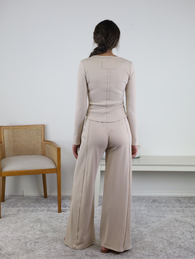 A woman stands barefoot on a gray rug, facing away. She wears the Statler Lounge Set, which includes a beige long-sleeve henley top and matching wide-leg pants. Beside her are a wooden chair with a cushion and a white console table placed against a plain wall.