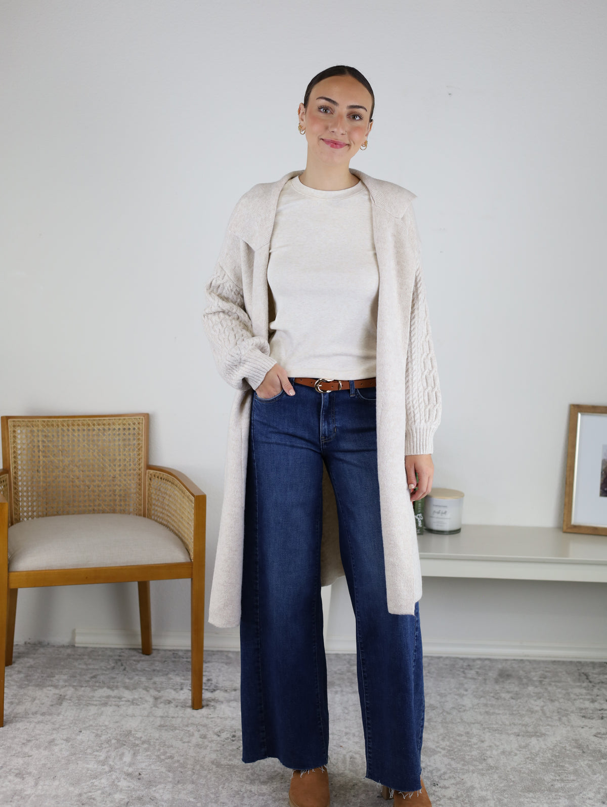 Wearing the Ashford Cable Knit Long Sleeve Knit Coat over a cream sweater and wide-leg jeans with a brown belt, the woman stands indoors near a wooden chair and bench adorned with framed art and a candle, set against a neutral wall.