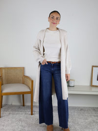 Wearing the Ashford Cable Knit Long Sleeve Knit Coat over a cream sweater and wide-leg jeans with a brown belt, the woman stands indoors near a wooden chair and bench adorned with framed art and a candle, set against a neutral wall.