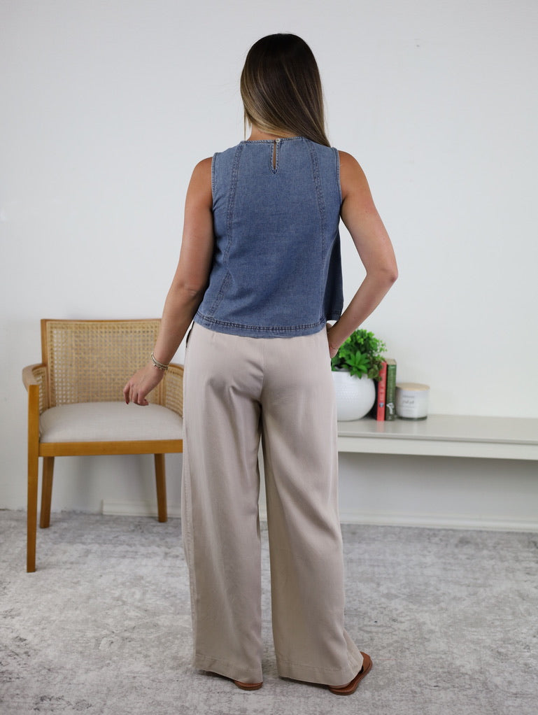 A woman with long hair stands facing away, wearing the Rebecca Seam Detail Sleeveless Denim Top – Final Sale and wide-leg beige pants. She is beside a light wood chair and a white shelf with books and decor on a light gray rug, highlighting the versatility of this summer blouse in an airy, modern setting.