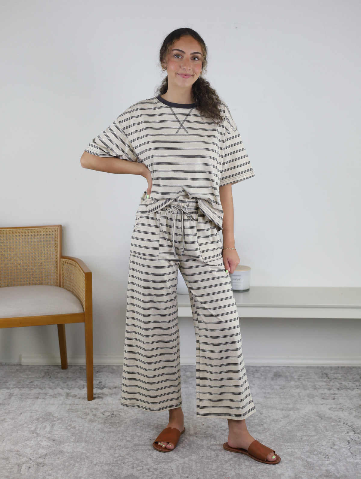 A woman stands indoors wearing the Lula Striped Pant Set, a cozy knit loungewear ensemble featuring wide-leg striped pants and a matching beige and gray top. She has one hand on her hip and is paired with brown sandals. A chair and bench can be seen in the background.