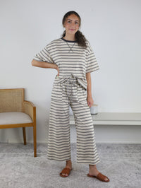 A woman stands indoors wearing the Lula Striped Pant Set, a cozy knit loungewear ensemble featuring wide-leg striped pants and a matching beige and gray top. She has one hand on her hip and is paired with brown sandals. A chair and bench can be seen in the background.