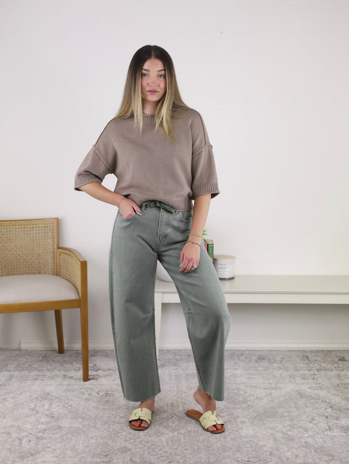 A woman stands indoors on a gray carpet, wearing a taupe sweater and light green Willy High Rise Barrel Jeans paired with sandals. She poses with one hand in her pocket beside a wooden chair and a table with jars.