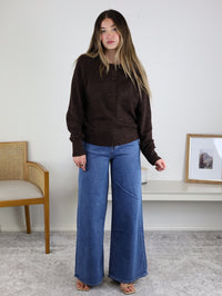 A woman with long hair wears the Theo Sweater and wide-leg jeans, standing indoors on a gray rug. A wooden chair and a low table with decor are behind her against a white wall.