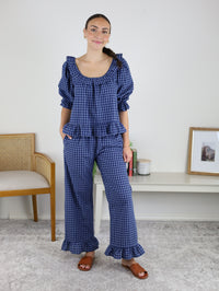A woman stands indoors, modeling the Barron Cotton Gingham Frill Set in blue—a coordinated ensemble featuring a frilled top and matching pants with ruffle accents. She smiles while wearing brown sandals, posed beside a beige chair and a white shelf adorned with decor and a framed photo—an ideal scene to highlight this stylish women’s loungewear.