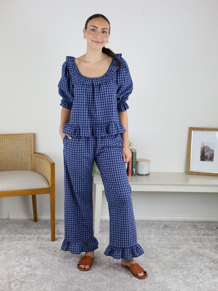 A woman stands indoors, modeling the Barron Cotton Gingham Frill Set in blue—a coordinated ensemble featuring a frilled top and matching pants with ruffle accents. She smiles while wearing brown sandals, posed beside a beige chair and a white shelf adorned with decor and a framed photo—an ideal scene to highlight this stylish women’s loungewear.