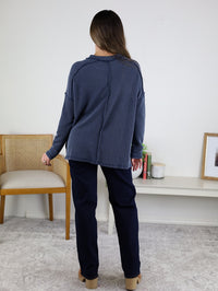 A person with long brown hair stands indoors facing away, wearing a navy ribbed knit Finley Sweater and dark pants, in a room with a beige chair, white bench, small plant, and framed artwork.