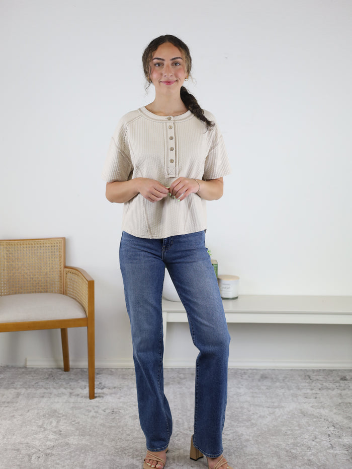 A woman with long dark hair in a loose braid stands indoors on a gray rug, smiling softly. She is wearing a light-colored Flynn Henley Knit Top paired with blue jeans. In the background, there is a wooden chair and a white bench adorned with decor.