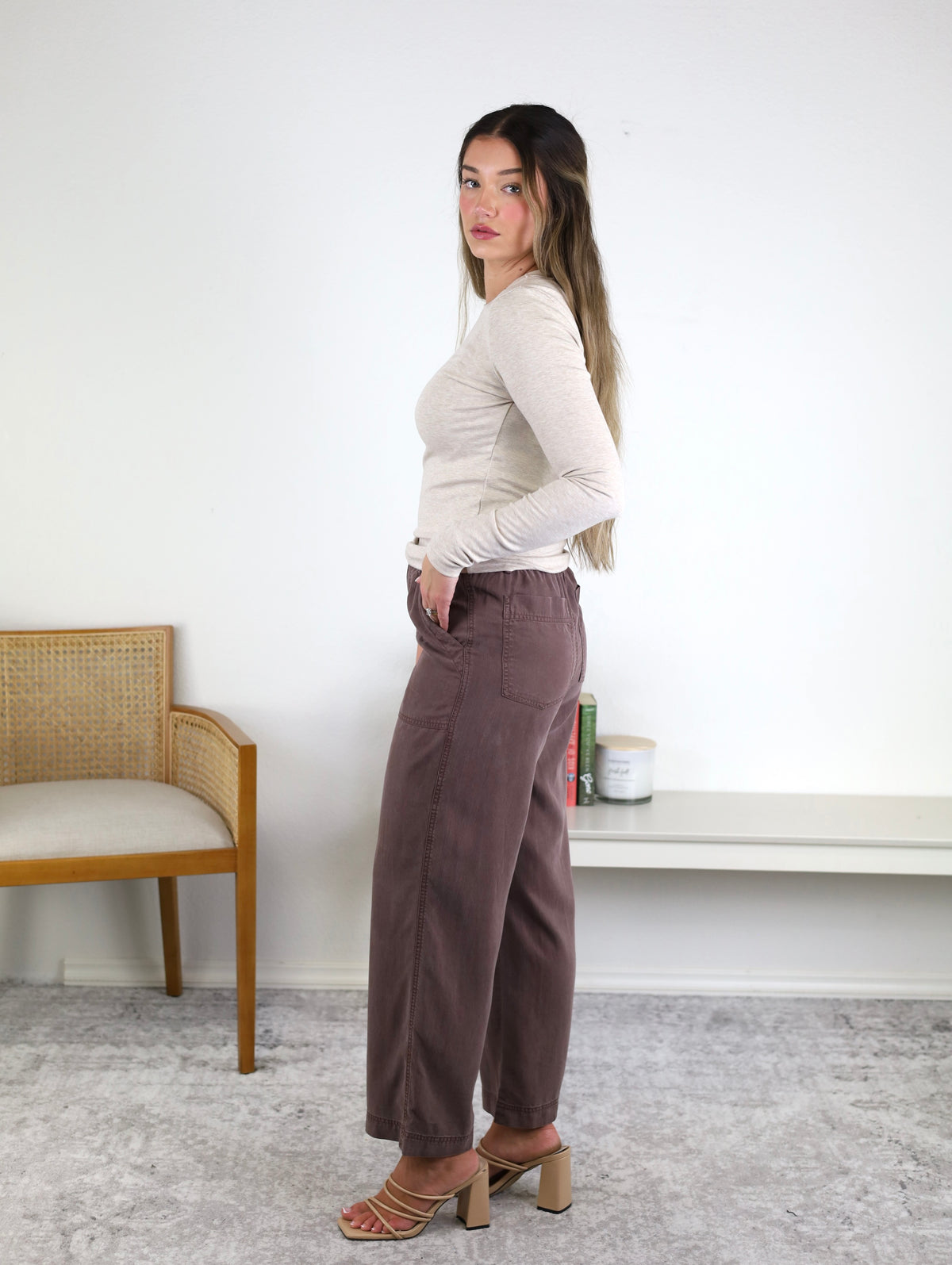 A woman with long hair stands sideways, wearing a beige long-sleeve top, heeled sandals, and the Marbella Wide Leg Tencel Pant in mauve with a drawstring elastic waist. A wooden chair and shelf with decor are visible in the background.