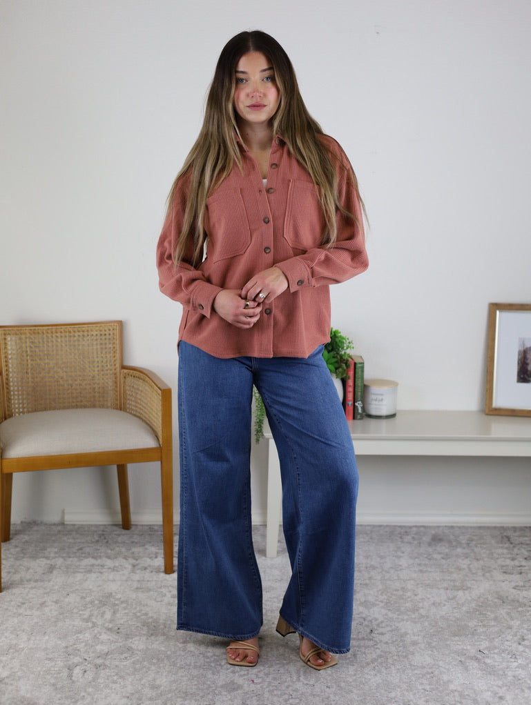 A woman with long hair wears the Larkin Rib Knit Button Up in dusty rose and wide-leg blue jeans, standing indoors on a gray carpet near a wooden chair and a white shelf.