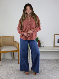 A woman with long hair wears the Larkin Rib Knit Button Up in dusty rose and wide-leg blue jeans, standing indoors on a gray carpet near a wooden chair and a white shelf.