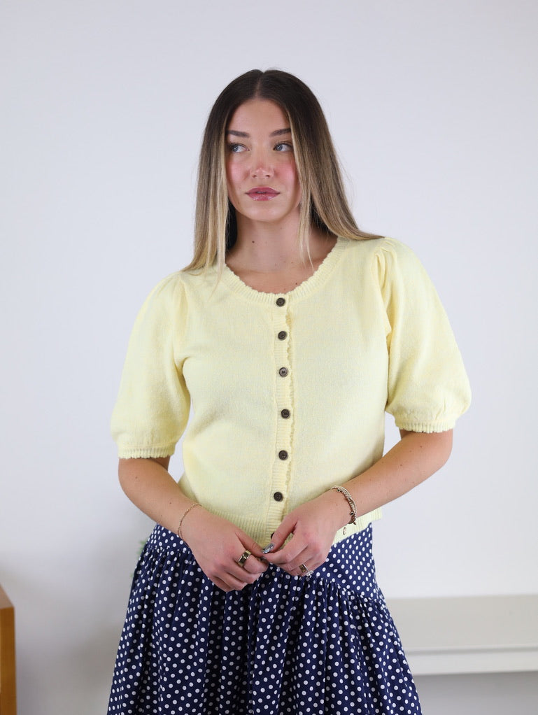 A woman with straight, long hair wears the Monica Short Sleeve Cardigan in light yellow, featuring metallic buttons, paired with a navy blue skirt with white polka dots. She is standing against a plain white background.
