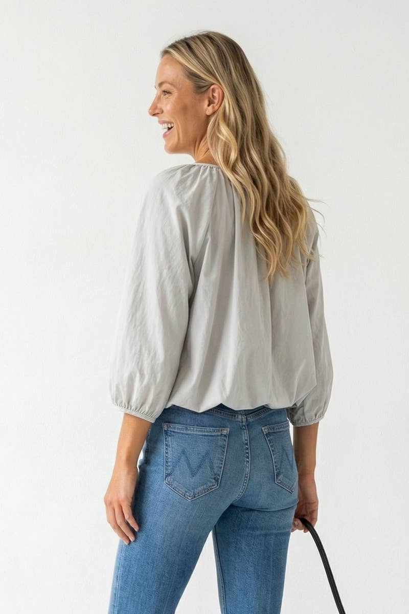 A woman wearing a Naked Zebra - Elastic Puff Sleeve V-neck Blouse in light gray with three-quarter length puff sleeves and blue jeans.