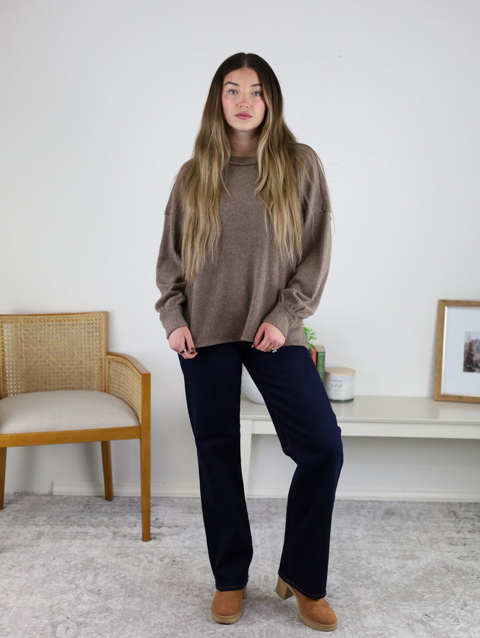 A woman with long hair wears the Lane Exposed Seam Top in brown, paired with dark blue pants and tan shoes. She stands indoors by a chair, near a table, plant, and framed picture against a white wall.