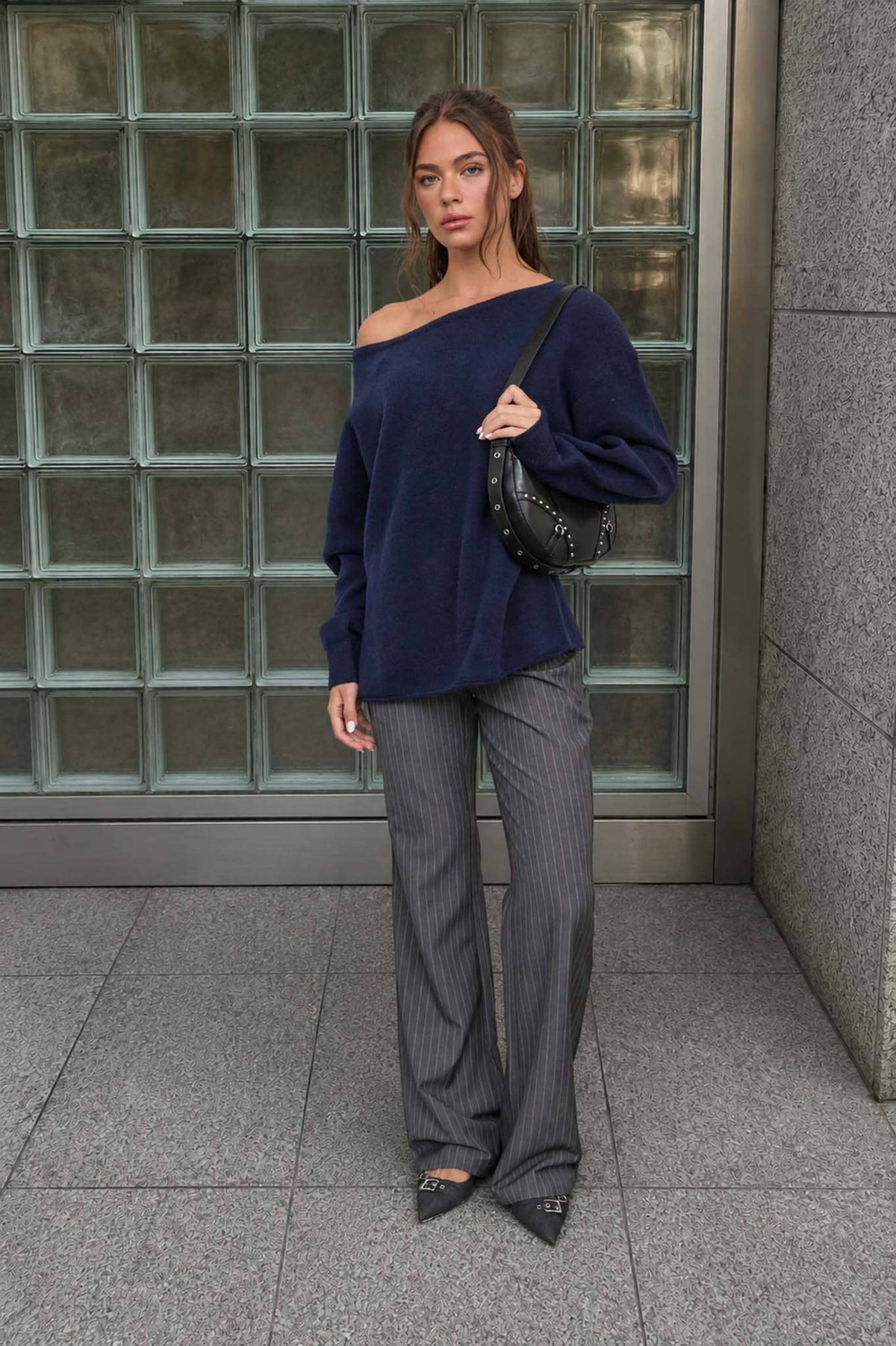 A woman stands in front of glass blocks, wearing The Workshop Studio’s Boat Neck Oversized Knit Pullover Sweater in Red Wood, paired with gray pinstripe pants, pointed shoes, and holding a black studded handbag.
