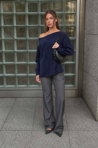 A woman stands in front of glass blocks, wearing The Workshop Studio’s Boat Neck Oversized Knit Pullover Sweater in Red Wood, paired with gray pinstripe pants, pointed shoes, and holding a black studded handbag.