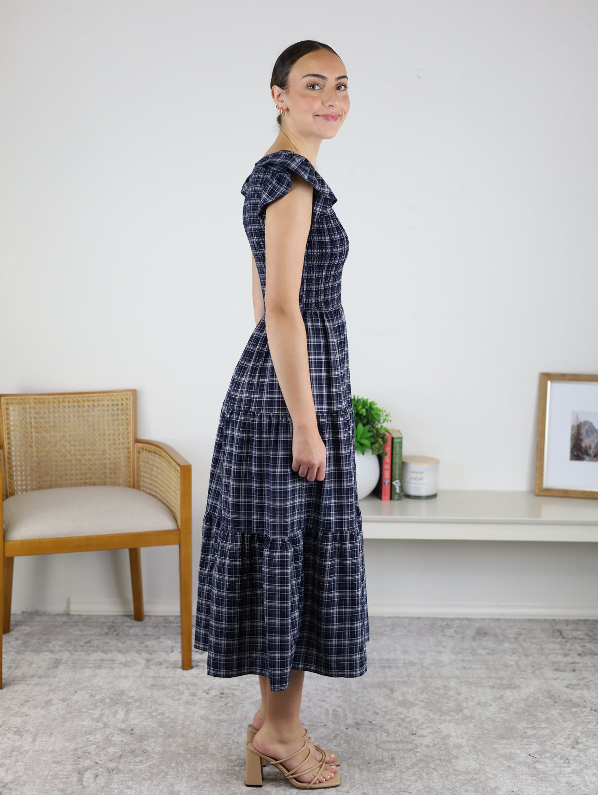 A woman in a Brynn Plaid V-Neck Tiered Midi Dress and beige heels stands sideways in a room with a chair, plant, books, and a framed photo on a white bench against a white wall—a perfect fall dress choice.