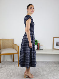 A woman in a Brynn Plaid V-Neck Tiered Midi Dress and beige heels stands sideways in a room with a chair, plant, books, and a framed photo on a white bench against a white wall—a perfect fall dress choice.