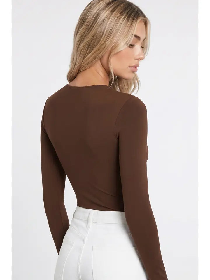 A woman with long blonde hair wears the High Neck LS Bodysuit in brown, paired with high-waisted white pants. She stands sideways and looks down against a plain background.