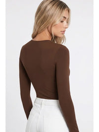 A woman with long blonde hair wears the High Neck LS Bodysuit in brown, paired with high-waisted white pants. She stands sideways and looks down against a plain background.