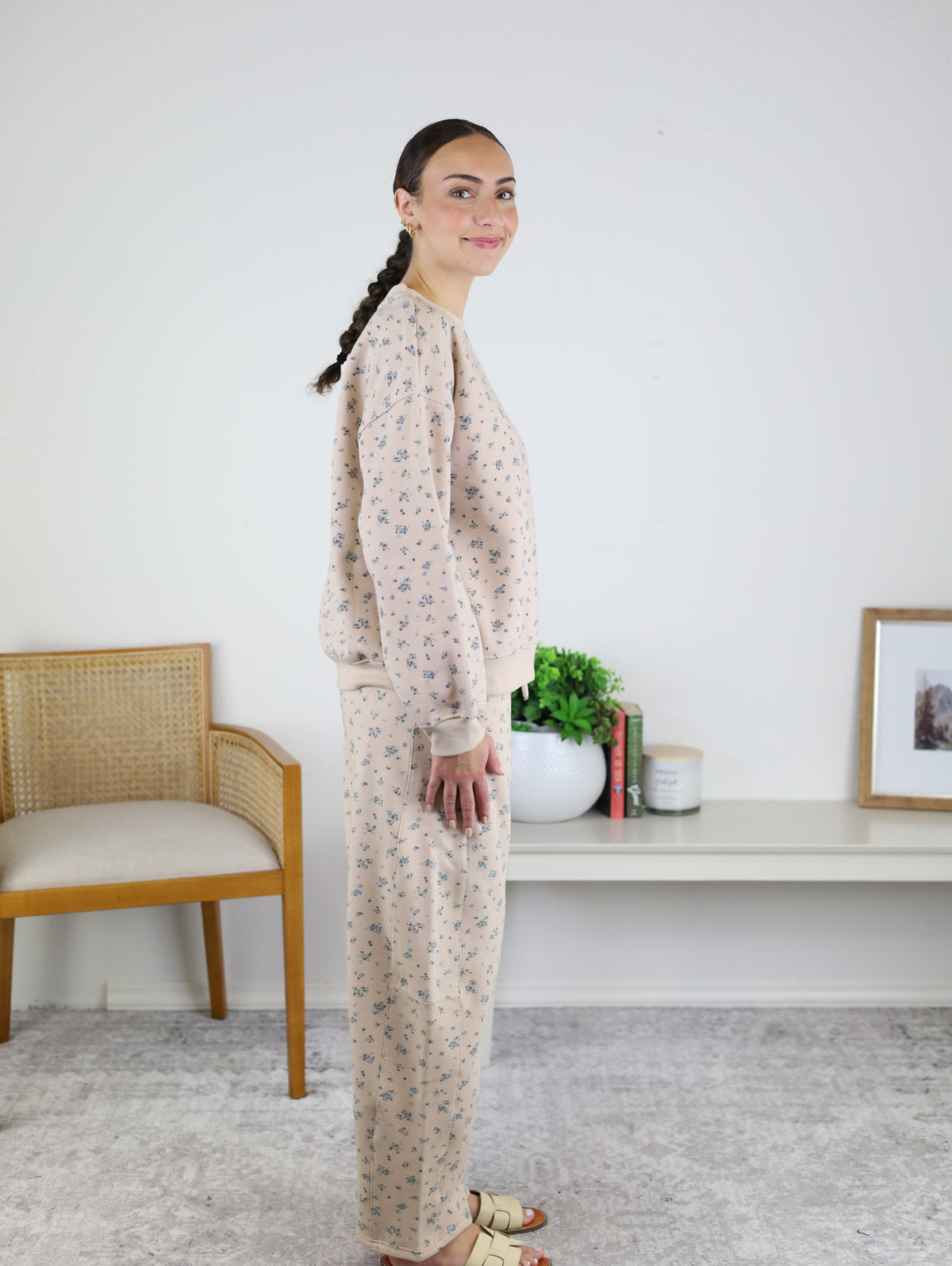 A woman stands in profile indoors, next to a wooden chair and a low shelf with plants and framed artwork, wearing the Journey Floral Boxy Crewneck Lounge Set—a women’s two-piece lounge set featuring a beige floral boxy crewneck with long sleeves and matching pants.