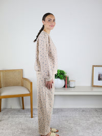 A woman stands in profile indoors, next to a wooden chair and a low shelf with plants and framed artwork, wearing the Journey Floral Boxy Crewneck Lounge Set—a women’s two-piece lounge set featuring a beige floral boxy crewneck with long sleeves and matching pants.