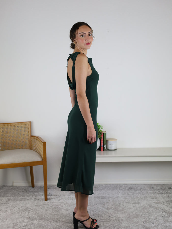 A woman wearing the Rudi Boat Neck Cowl Back Midi Dress with black heels stands sideways in a minimalist room featuring a chair, books, and small decor on a white shelf.