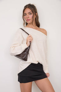 A woman stands against a plain background, holding a dark brown shoulder bag and wearing The Workshop Studio - BOAT NECK OVERSIZED KNIT PULLOVER SWEATER in Oatmeal with a black skirt, looking at the camera with a neutral expression.