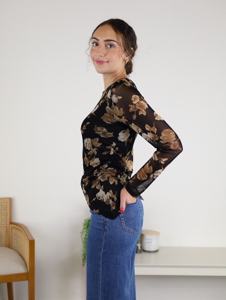 A woman with brown hair in a bun is wearing the Nightfall Rose Floral Mesh Asymmetrical Top, featuring black sheer mesh sleeves and a floral design, paired with blue jeans. She stands sideways and smiles in a minimal room decorated with a chair, a plant, and a candle on a white shelf.