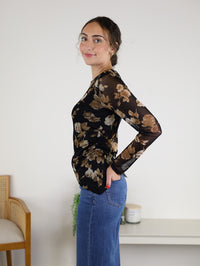 A woman with brown hair in a bun is wearing the Nightfall Rose Floral Mesh Asymmetrical Top, featuring black sheer mesh sleeves and a floral design, paired with blue jeans. She stands sideways and smiles in a minimal room decorated with a chair, a plant, and a candle on a white shelf.