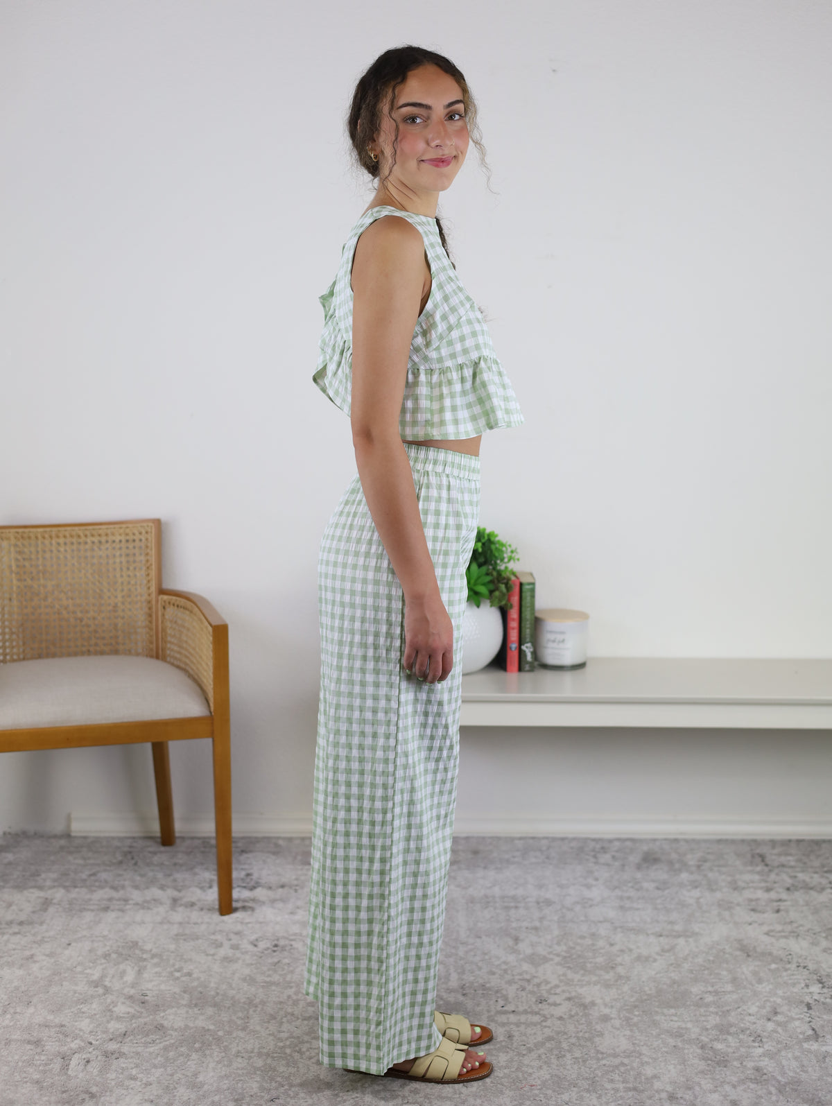 A woman stands sideways, smiling slightly, wearing the Myra Gingham Print Ruffled Set in sage—a sleeveless crop top paired with matching high-waisted wide-leg pants. In the background are a chair, a low white shelf, potted plants, and stacked books.