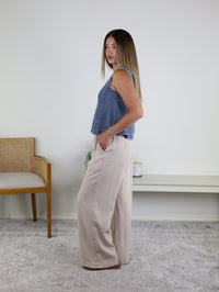 A woman stands sideways in a room, wearing the Rebecca Seam Detail Sleeveless Denim Top—styled as a versatile summer blouse—paired with wide-legged beige pants. She has straight, long hair and rests one hand in her pocket. In the background are a wooden chair and a shelf.