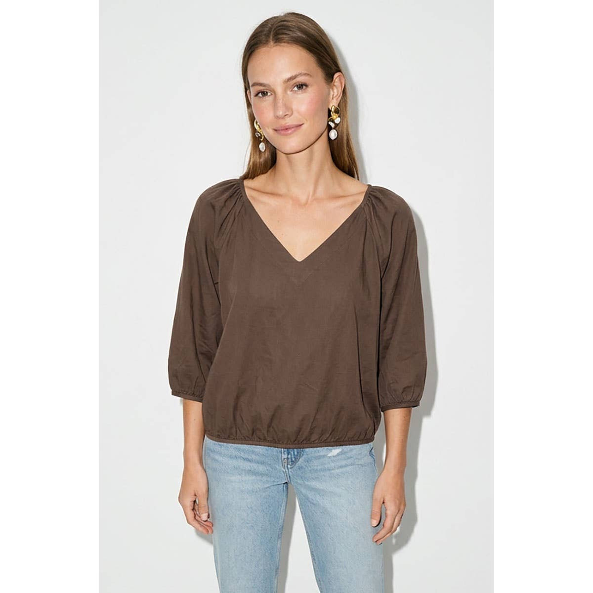 A woman wears a Naked Zebra - Elastic Puff Sleeve V-neck Blouse: Burgundy / L featuring three-quarter length puff sleeves and an elasticized hem, paired with light-wash blue jeans.
