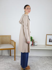 A woman stands sideways indoors, wearing the Pinch Me Long Cardigan over blue jeans and tan boots. She’s posed in front of a white wall near a wooden chair and a low table with books, a plant, and a framed picture—the perfect look for cozy layering on brisk days.