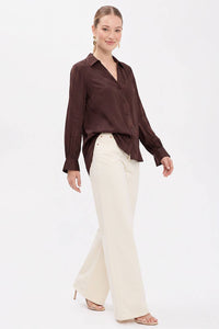 A woman wearing a Naked Zebra - Neck Shirt With Open Seam Sleeve: Black / L in brown tucked into cream-colored wide-leg trousers.