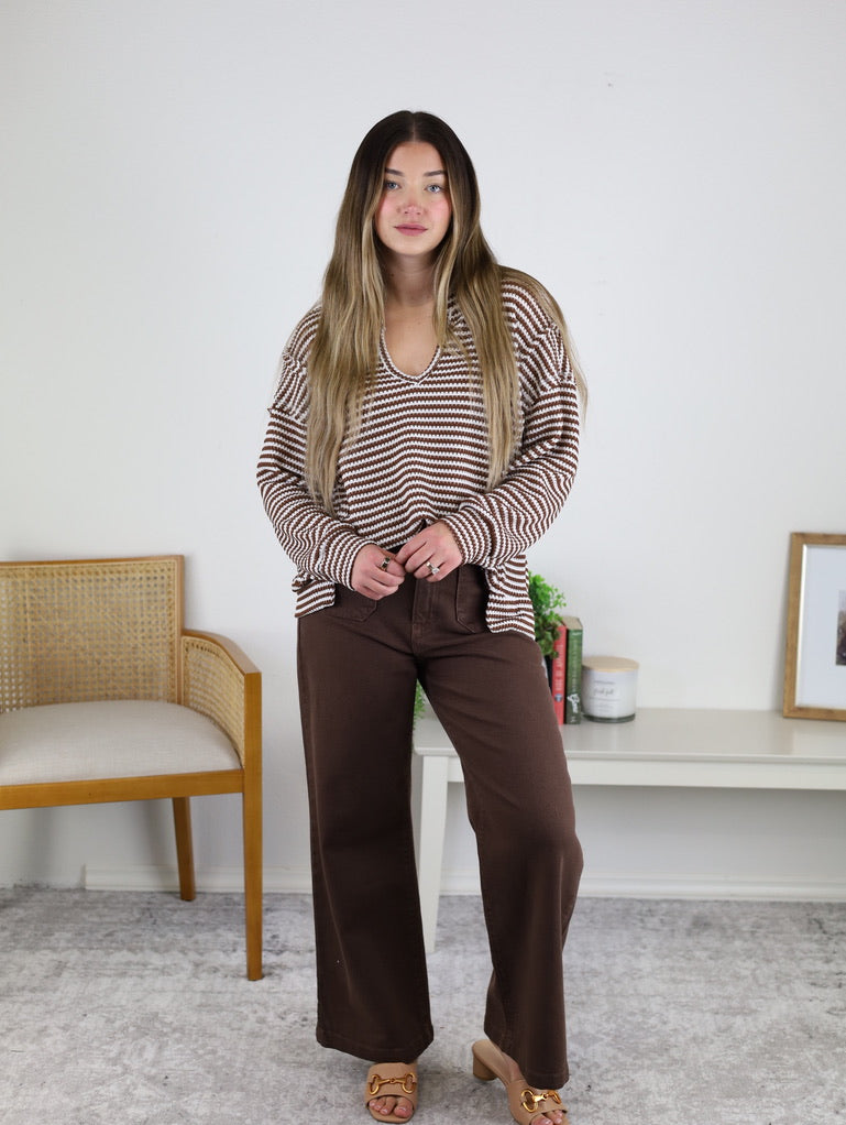 A woman with long hair wears the Lennon Stripe Knit Top and brown pants, standing indoors near a wooden chair and a white console table with books, a candle, and framed art.