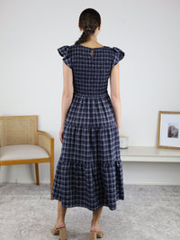 A woman stands indoors facing away, wearing the Brynn Plaid V-Neck Tiered Midi Dress—a navy and white plaid, short-sleeve, tiered dress with ruffle accents perfect for fall. Her hair is tied back and she wears beige sandals. A chair and a framed photo appear in the background.