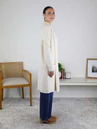 A woman stands in profile indoors, smiling with her hair tied back, wearing the Parkland Long Cardigan in cream over blue jeans and tan shoes. She poses next to a chair and a shelf decorated with various items.