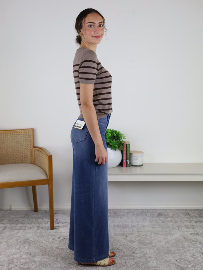Wearing the Rachelle Low Rise Denim, a woman in a striped shirt stands sideways and smiles. She is inside a room with a chair, a white shelf filled with books, and a plant in the background.