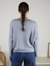 A person with a single braid is wearing the Maren Striped Cardigan, styled in a relaxed fit with blue and white stripes, along with blue jeans, standing with their back to the camera in a simple indoor setting.