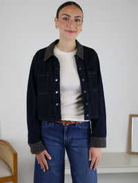 A woman with dark hair in a bun wears a cream shirt and the Autumn Leopard Collar Denim Shacket, styled effortlessly with blue jeans. She is smiling and standing indoors beside a bench and a small white table topped with a framed picture.