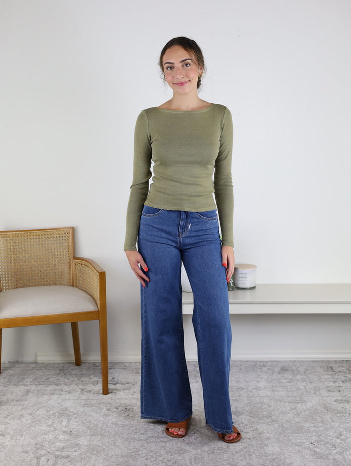 A woman stands indoors on a gray rug, wearing the Clove Fitted Rib Knit Boat Neck Top in green, blue wide-leg jeans, and brown sandals. A wooden chair and a white bench with a small candle are visible in the background.