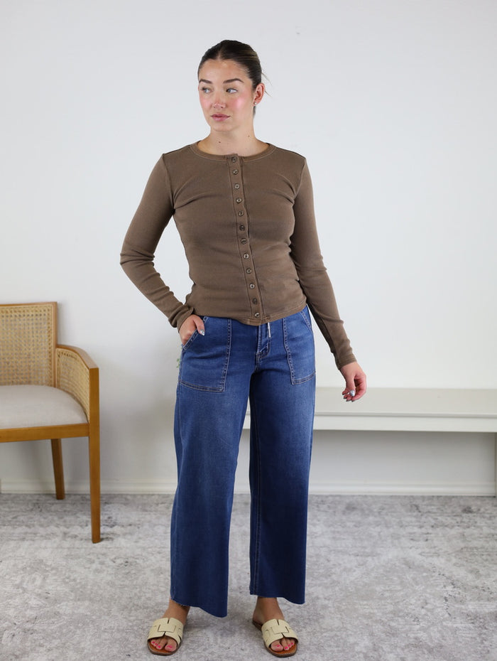 A woman stands indoors wearing the Arden Button Front Top, paired with wide-leg blue jeans and beige sandals. Her hair is tied back as she looks to the side, with a wooden chair and a white bench in the background.