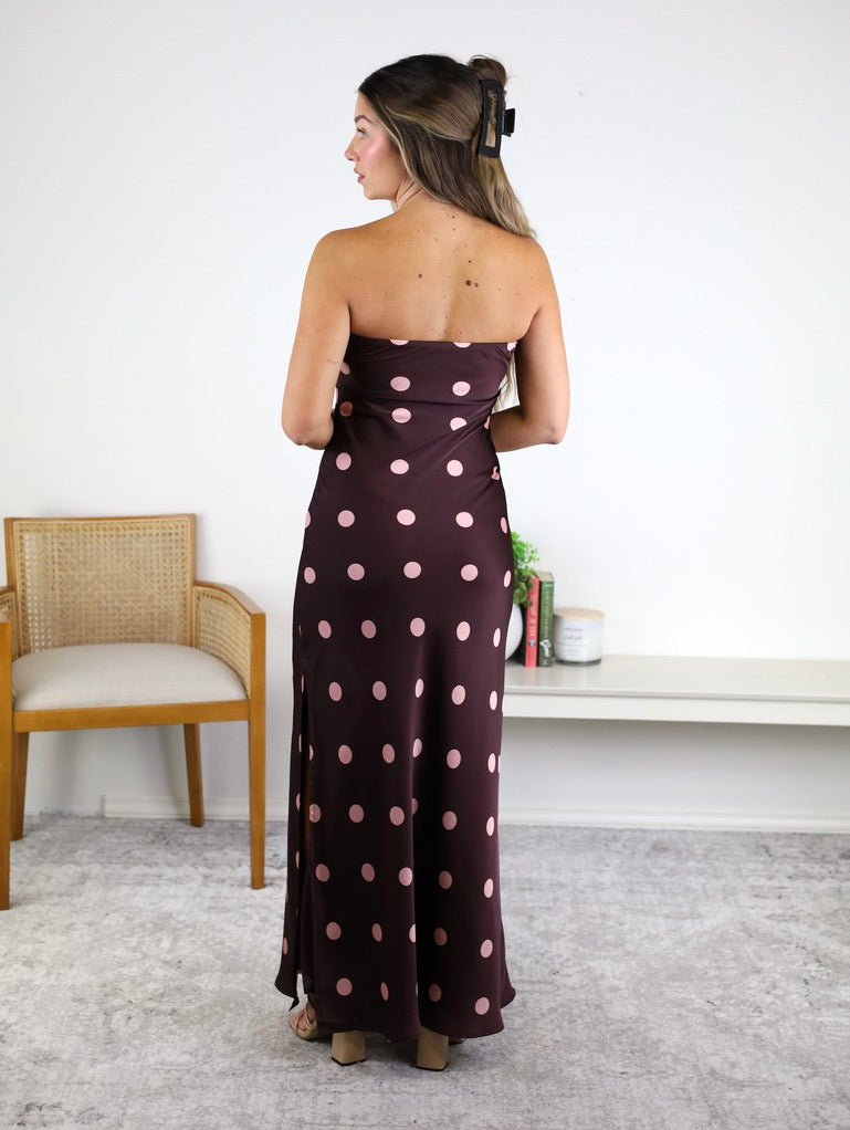 A woman with long hair clipped back stands with her back to the camera, wearing the Polka Dot Satin Strapless Maxi Dress in dark brown with a large mauve dot print. In the background, there is a chair and a white bench adorned with decor.