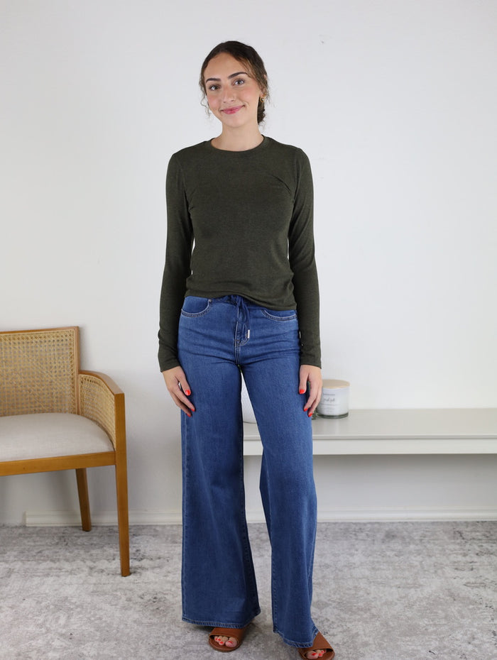 A woman stands indoors on a gray rug, wearing the Josette Ribbed Classic Crewneck in dark green and wide-leg blue jeans. There is a wooden chair and a white shelf with a candle behind her.