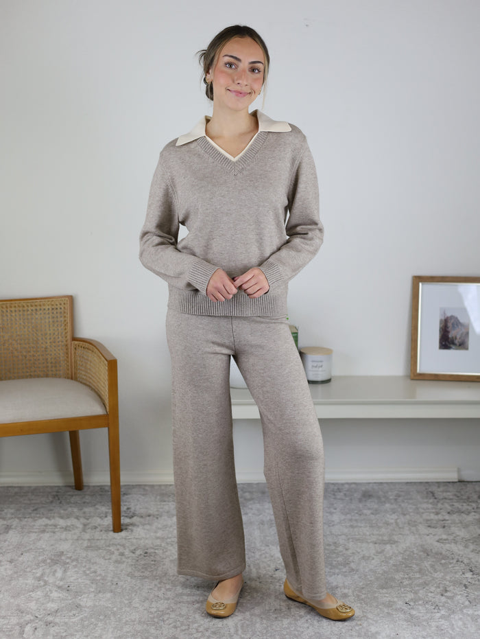 A person wearing the Rebecca Sweater/Pant Set, a cozy loungewear outfit consisting of a beige V-neck sweater with a contrasting cream collar and matching wide-leg trousers.