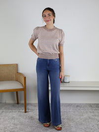 A woman is standing indoors wearing the Ace Puff Striped Sweater—a beige, striped, lightweight knit with short sleeves—paired with wide-leg jeans and sandals. She is smiling with one hand in her pocket, with a chair and shelf visible in the background.