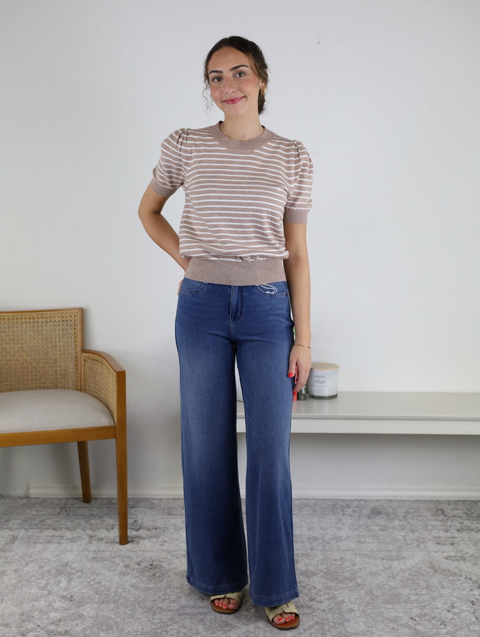 A woman is standing indoors wearing the Ace Puff Striped Sweater—a beige, striped, lightweight knit with short sleeves—paired with wide-leg jeans and sandals. She is smiling with one hand in her pocket, with a chair and shelf visible in the background.