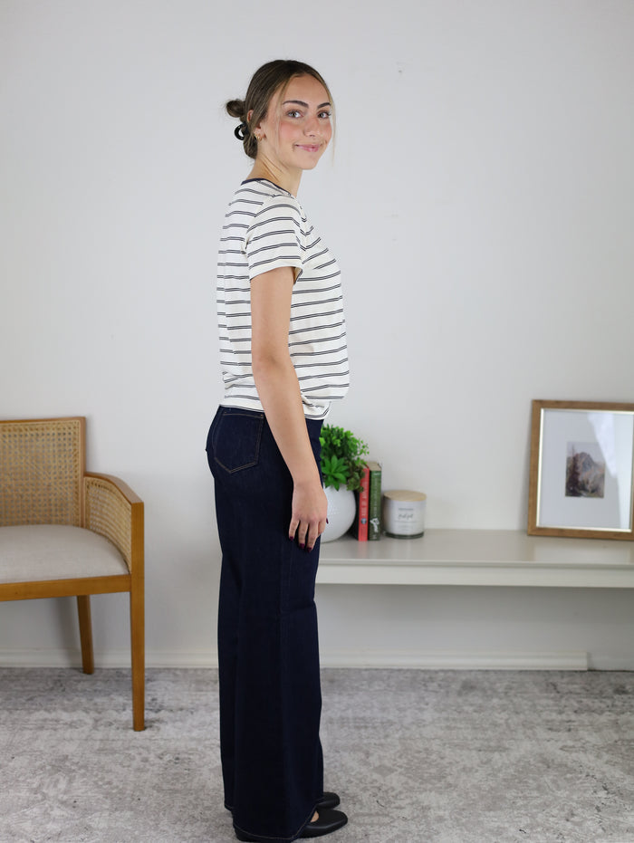 A woman in profile wears a black and white striped Asher Tee and dark blue wide-leg jeans.