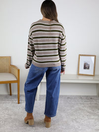 A woman stands indoors facing away, wearing a striped sweater, tan shoes, and the Kate Double Pocket Barrel Jean. She is near a white table with a framed photo and a wooden chair on a light carpeted floor.